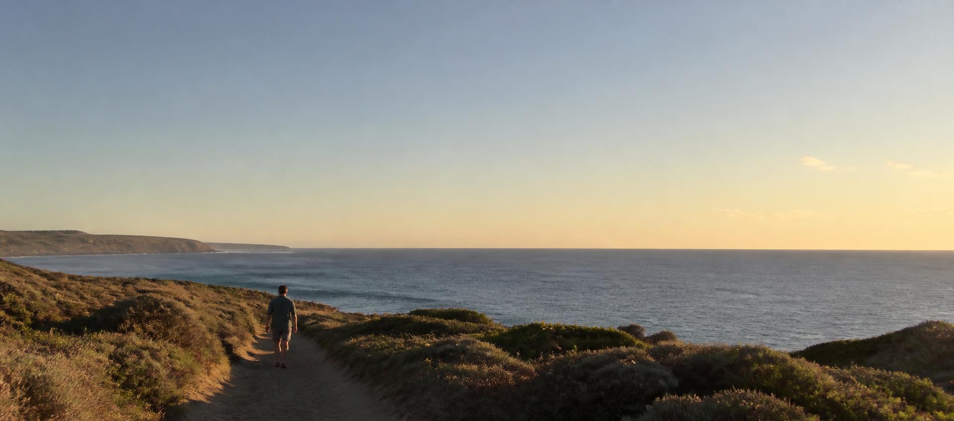 A person walking a sandy coastal path toward an open ocean horizon in late afternoon light.